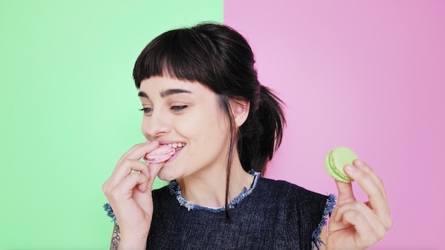 A woman eating a macaroon on a pink and green background. 